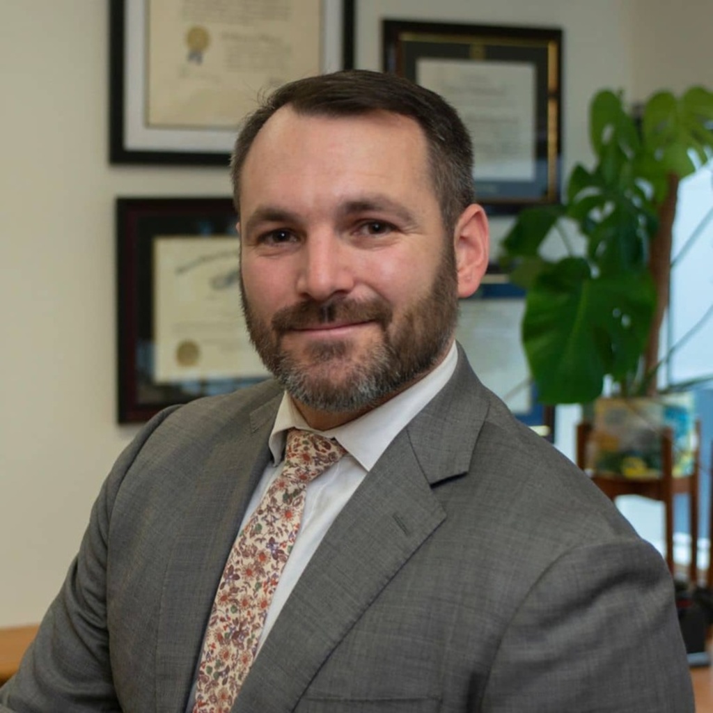 A headshot of Mihailis Diamantis with framed law degrees and a plant in the background